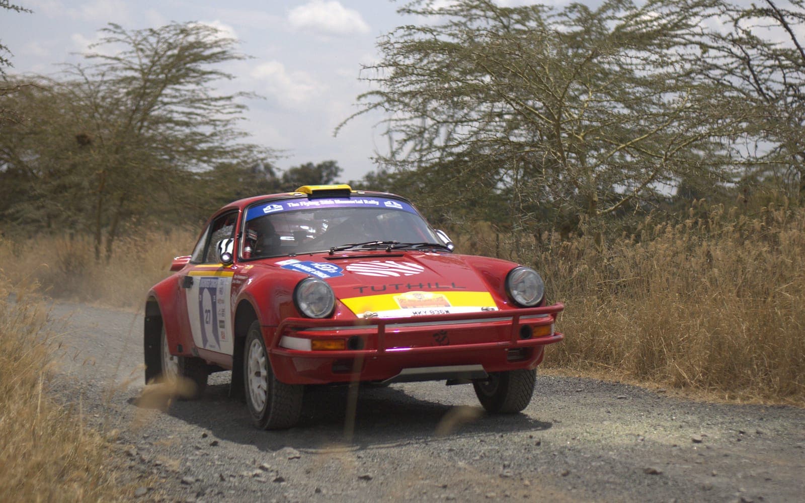 The competition car on murram between the thorn trees, dust rising behind